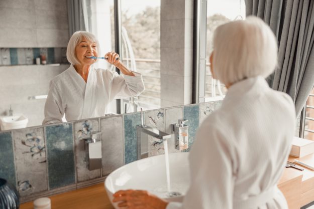 Lovely old lady brushing teeth in bathroom From Bathing To Grooming: The Importance Of Personal Care In Florida Nursing Homes