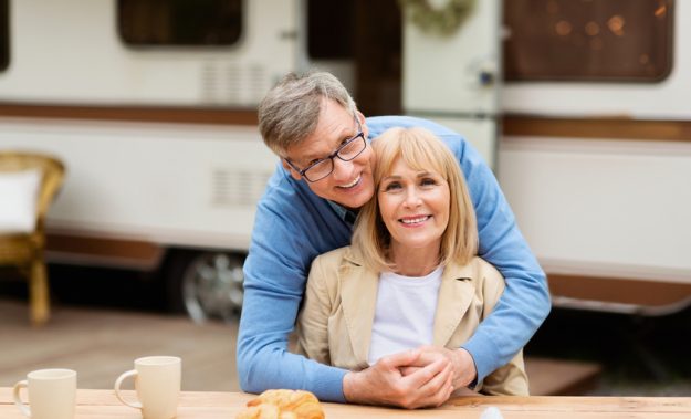 Smiling mature man hugging his beautiful wife in front of camping vehicle outdoors Smiling mature man hugging his beautiful wife in front of camping vehicle outdoors