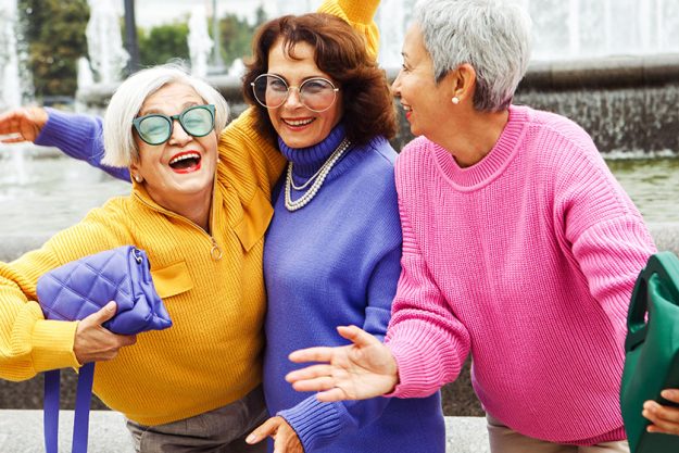 Three happy cheerful pensioner girlfriends in bright sweaters and sunglasses walk together in the city on an autumn day. Three happy cheerful pensioner girlfriends in bright sweaters and sunglasses walk together in the city on an autumn day.